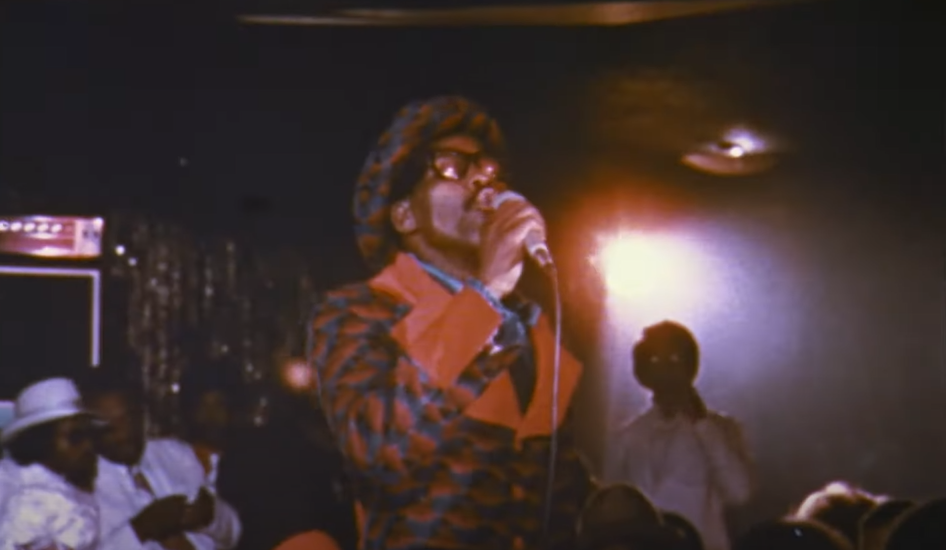 Man in coat and sunglasses singing with a mic in a dark room