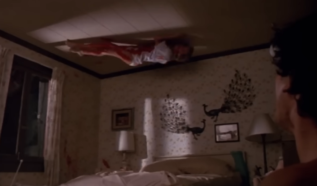 bloodied woman on the ceiling in the room, a man in the corner stands and observes