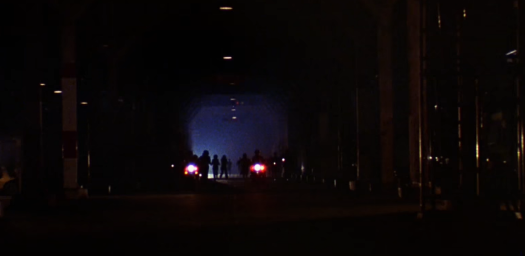 A dark street with a bright light at the end and silhouettes of people standing there